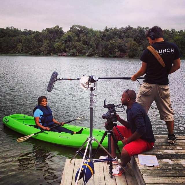 Two camera men film an African American woman preparing to kayak on a lake