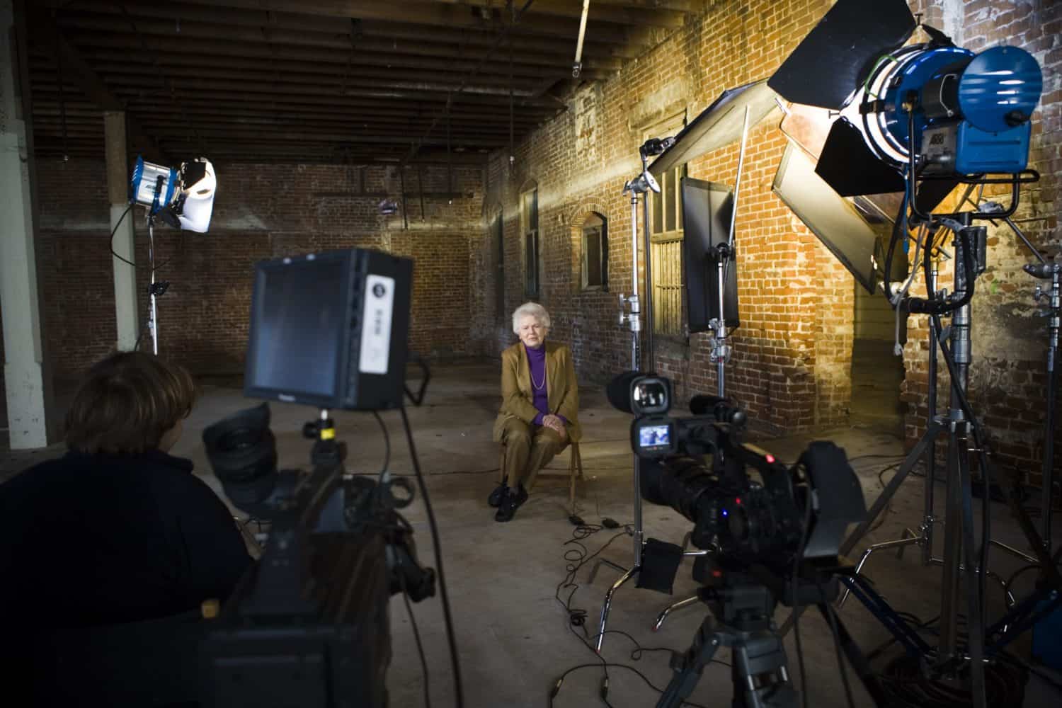 A camera crew films an old woman in a brick warehouse